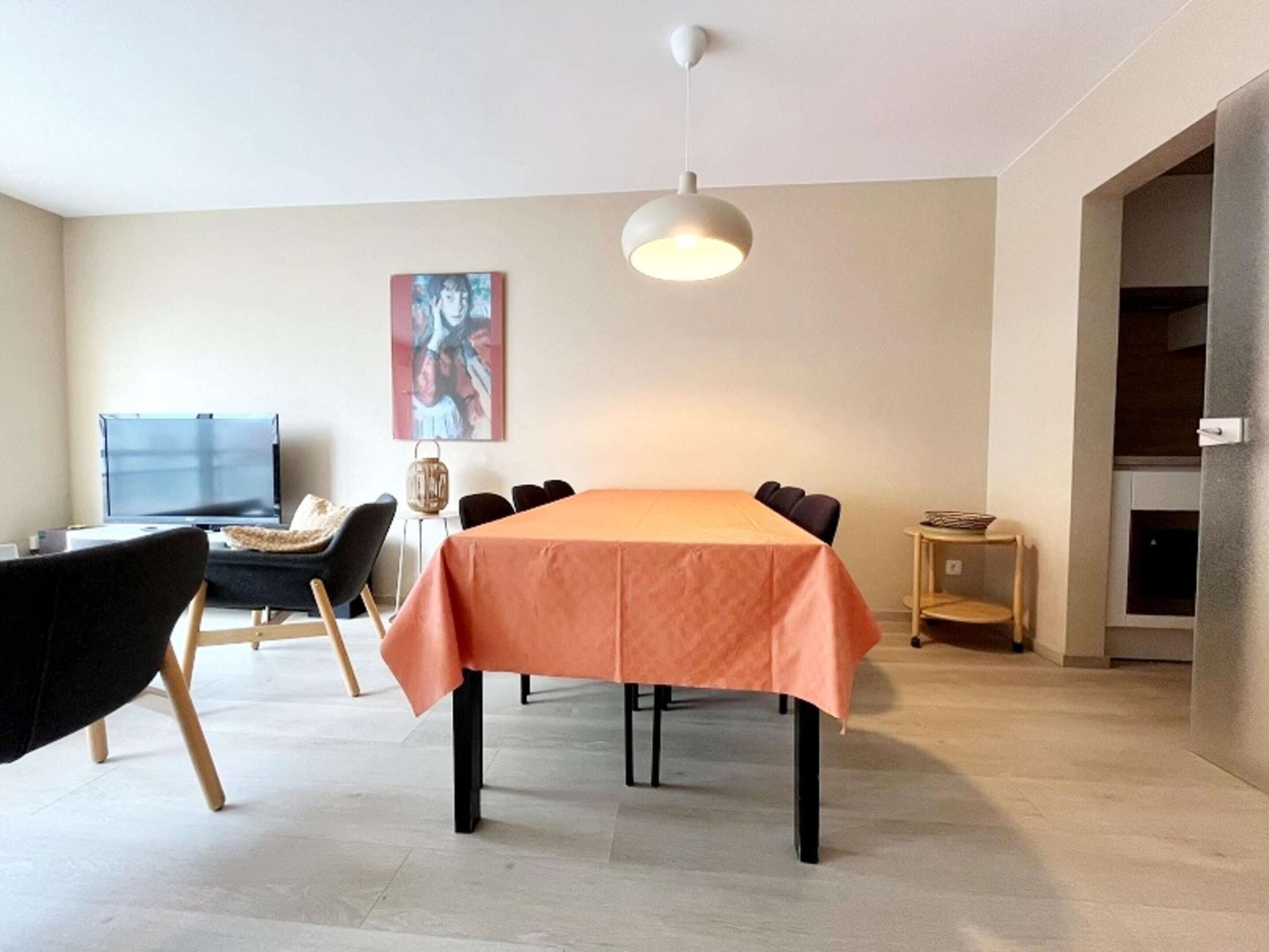 Un'area pranzo moderna con un tavolo ricoperto da una tovaglia color pesca, sedie nere, un lampadario, un televisore e un quadro appeso al muro.