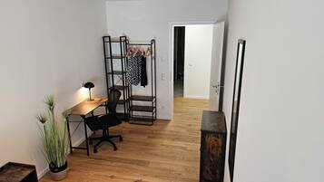 A compact room with a wooden desk, a black office chair, a small potted plant, a black metal shelf with clothes, and a wooden door.