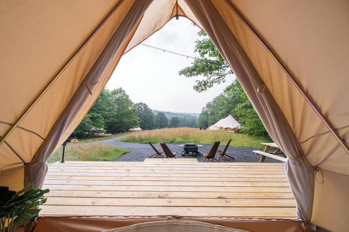 Ein Zelt mit Holzterrasse, Feuerstelle und Stühlen, die auf eine Wiese mit Bäumen und Hügeln im Hintergrund ausgerichtet sind.