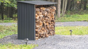 A metal shed with a stack of firewood inside, a small lamp on the ground, and a gravel path in a wooded area. - Firefly Ridge (Great Cacapon)