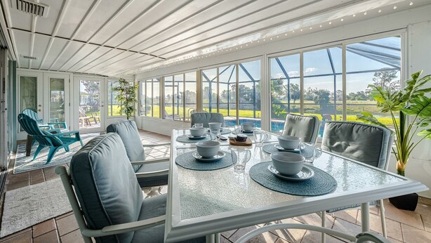 Une véranda avec une table en verre dressée pour les repas, entourée de sièges confortables et de grandes fenêtres donnant sur un jardin.