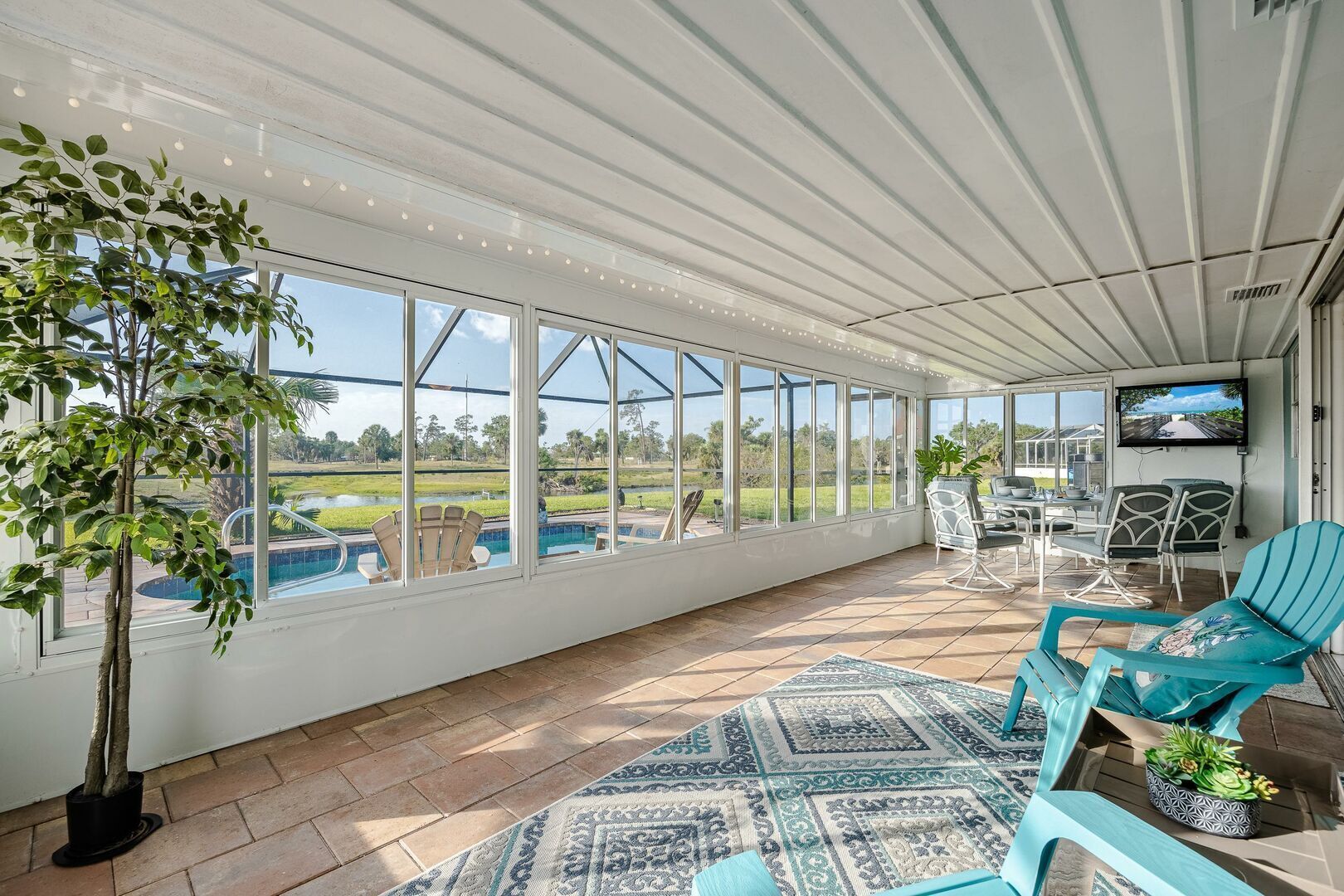 A covered patio with a view of a swimming pool, outdoor furniture, and a television.