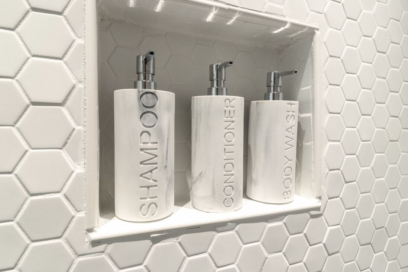 Une étagère de salle de bain avec trois flacons blancs étiquetés « SHAMPOO », « APRÈS-SHAMPOO » et « GEL DOUCHE ».