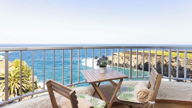 Un balcon avec une table en bois et deux chaises, donnant sur une côte rocheuse et l’océan.