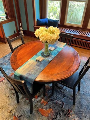 A round wooden dining table with a vase of yellow flowers, black chairs, and a blue and green patterned runner. - CRAFTSMAN's MAIN FLOOR 2 bd rm- chef's kit- deck (Willmar)