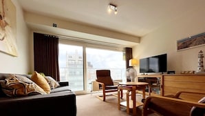 Camera d'albergo con divano, poltrone, televisione, scrivania e un'ampia finestra vista città.