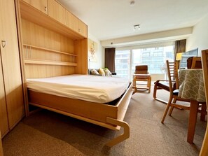 Habitación de hotel con cama, armario de madera, escritorio con silla y ventana con vista a edificios.