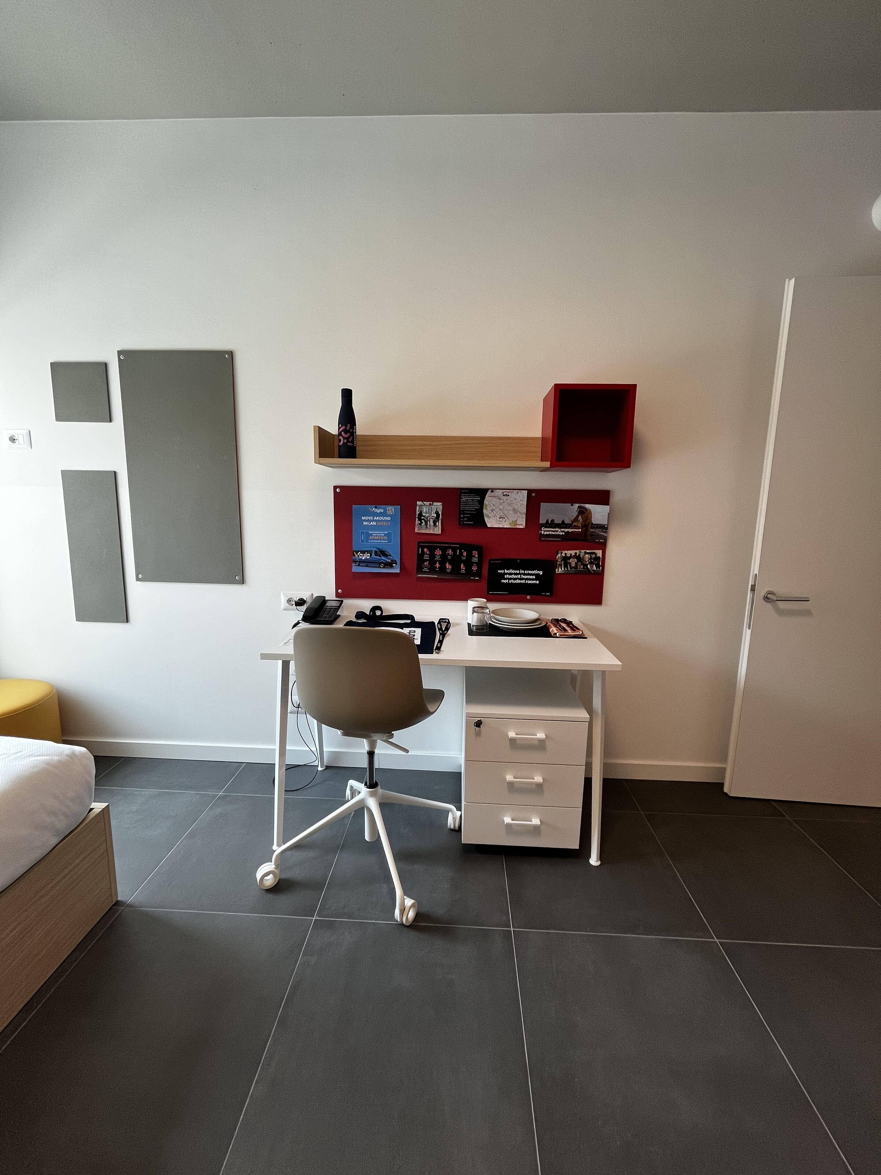 Un dormitorio moderno con una cama, un escritorio y una silla. Hay un estante rojo con varios objetos y un mapa en la pared.