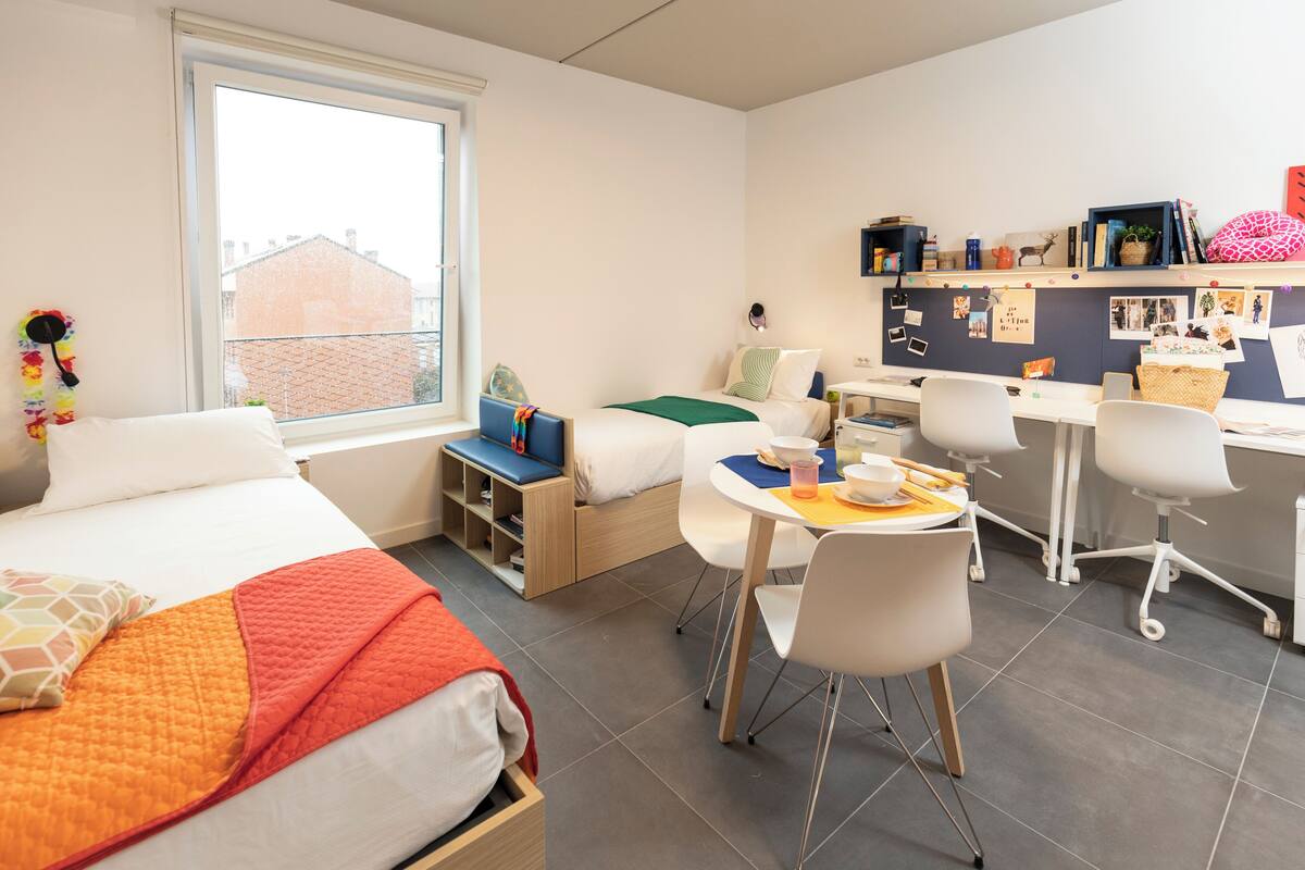 A modern hotel room with a bed, a desk, a chair, a small seating area, and a window with a view of buildings.