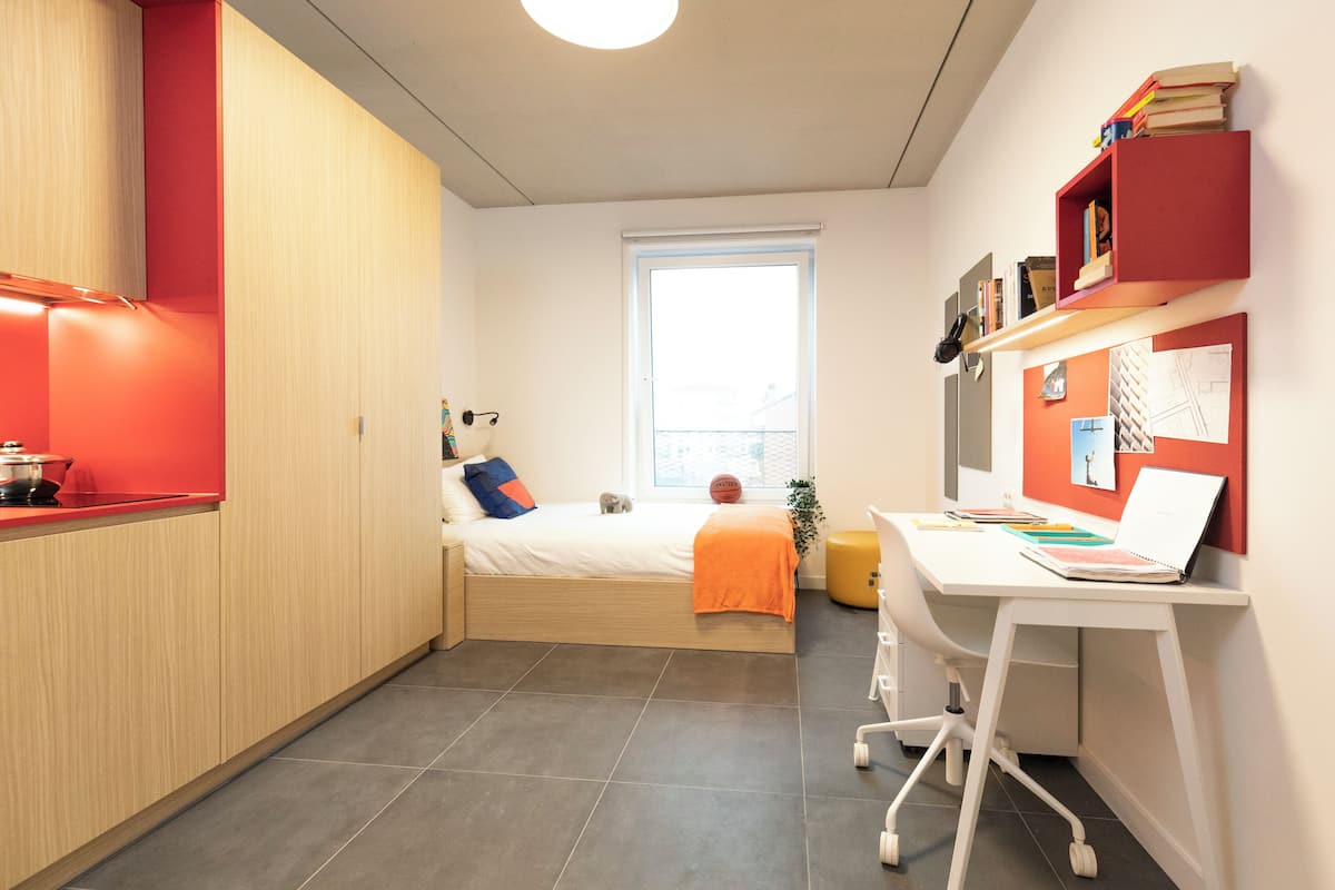 A compact room with a bed, a desk, a red shelf, and a window.