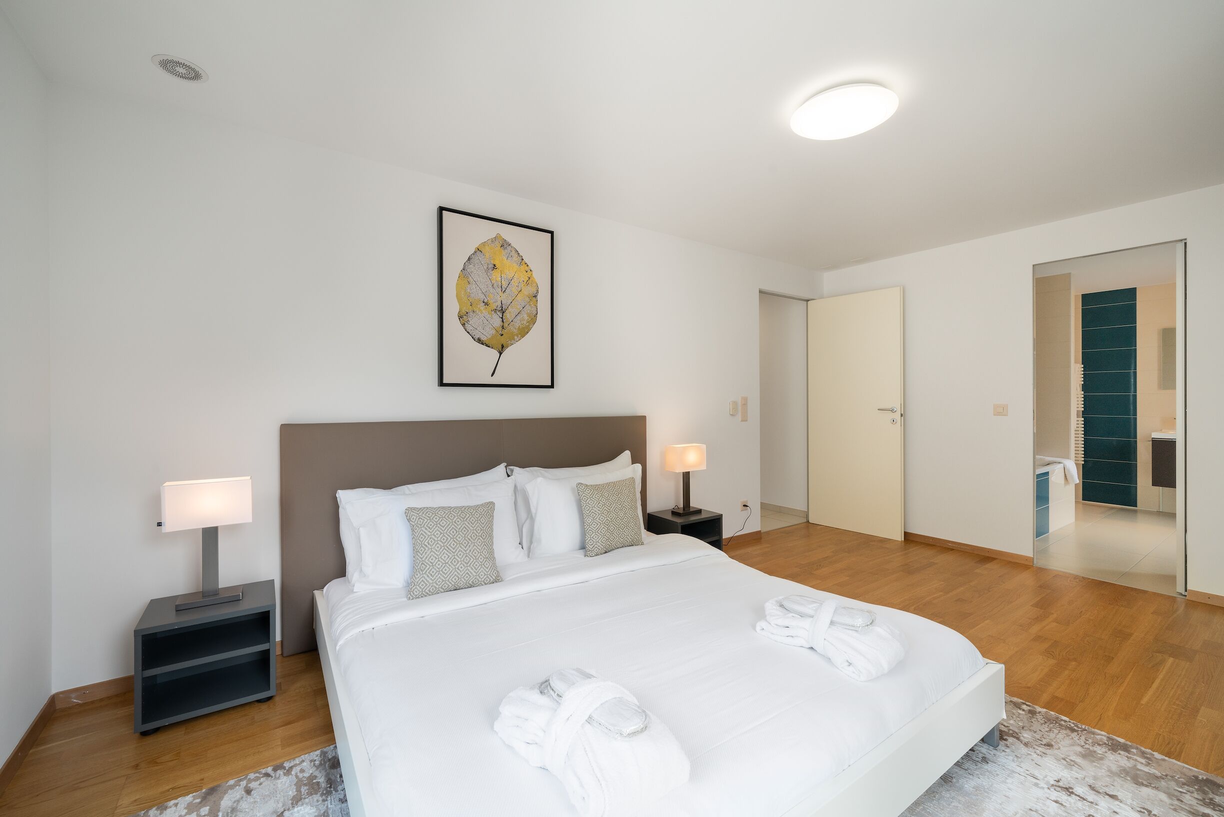 A modern hotel room with a large bed, bedside lamps, a framed leaf artwork, and a bathroom visible through an open door.