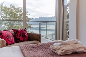 A room with a bed, a sofa with patterned cushions, a view of a lake and mountains, and a balcony with a glass railing.