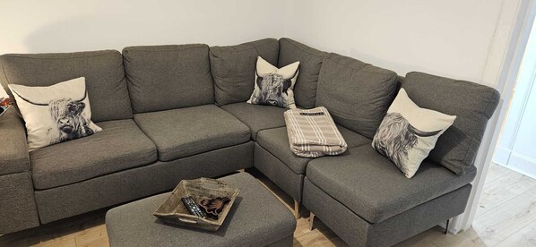 A grey sectional sofa with a small ottoman, two decorative pillows, and a remote control on the ottoman. - Modern, 2 bedroom, pet-friendly studio, close to Scotlands best golf courses! (Cupar)