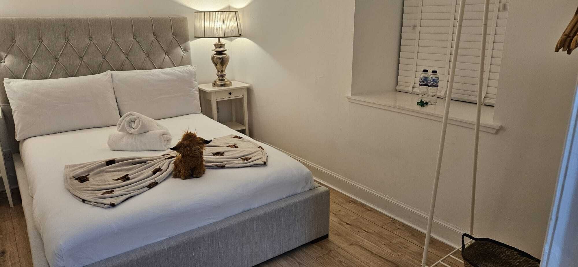 En pænt redt seng med et blødt tæppe, en pude og et sammenrullet håndklæde. Der er et natbord med en lampe og en vandflaske på.