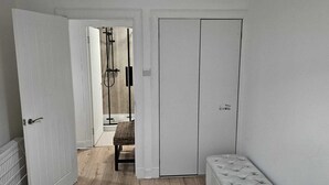 Um corredor estreito com uma porta branca à direita, um radiador à esquerda e um banco de vime no centro.