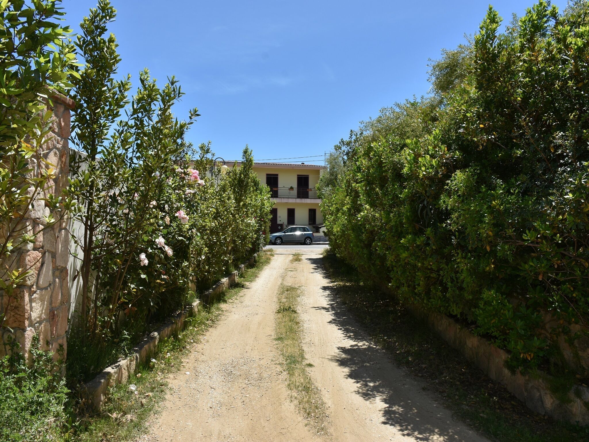 Un chemin de terre mène à un bâtiment doté d’un balcon, qui est entouré de verdure et où se trouve une voiture garée.