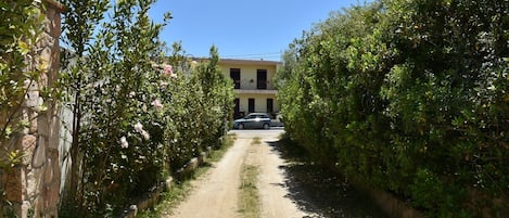 Un chemin de terre mène à un bâtiment doté d’un balcon, qui est entouré de verdure et où se trouve une voiture garée.