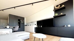 Un soggiorno moderno con un divano grigio, un piccolo tavolo bianco e un angolo cottura con mobili bianchi e una parete di accent in nero.