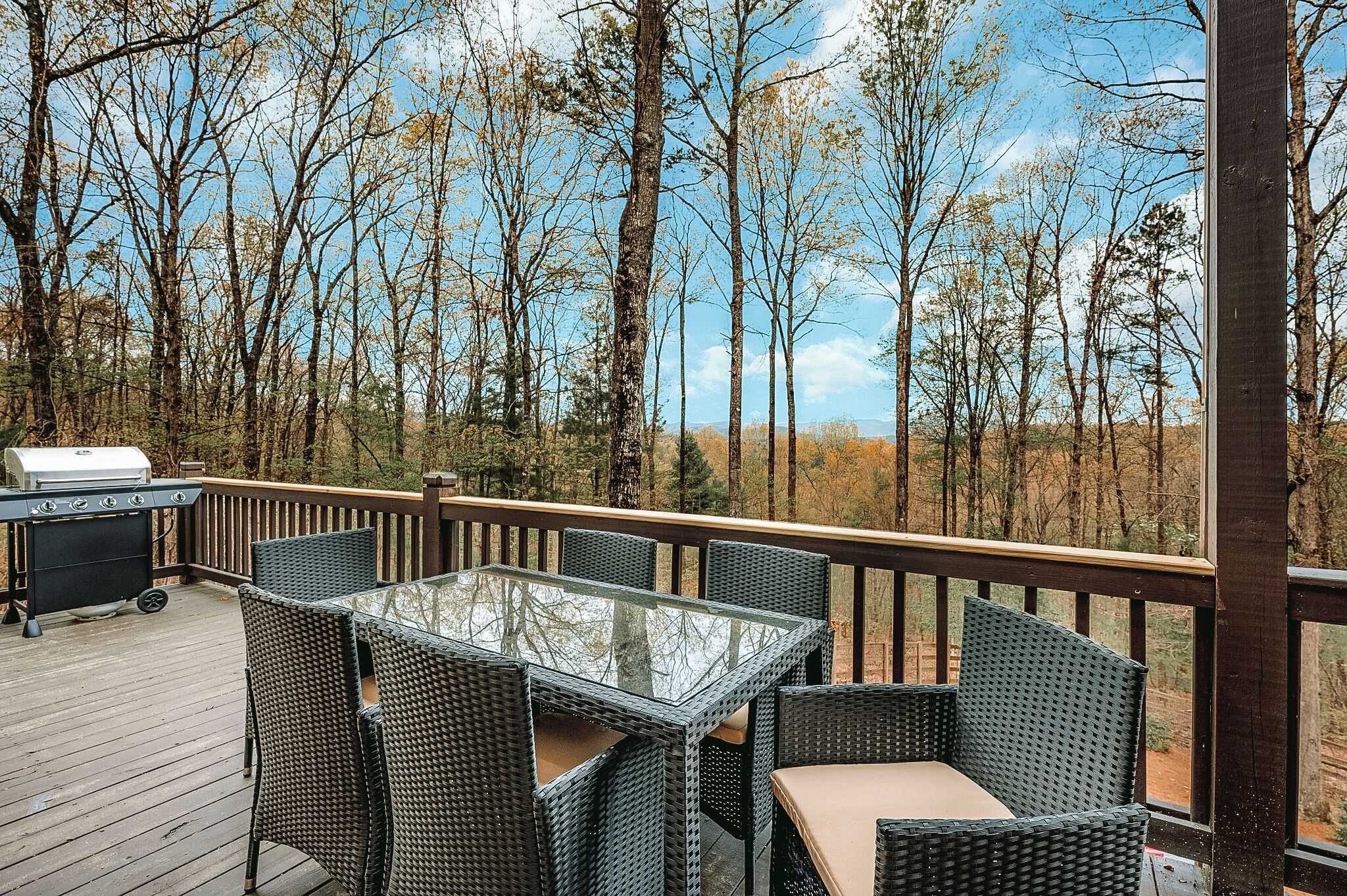 A deck with a glass-top table and wicker chairs, surrounded by a wooden railing, overlooking a forested area.