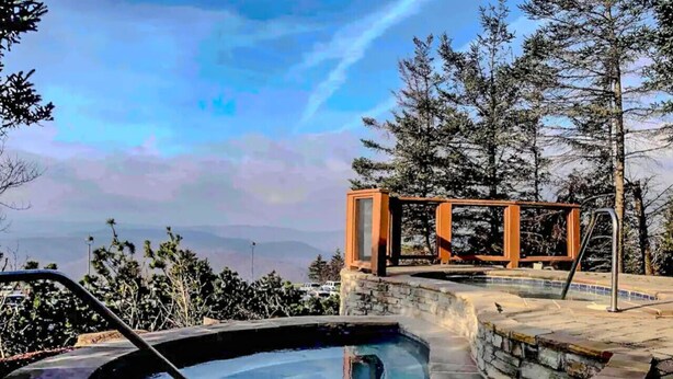 A hot tub with a stone edge, surrounded by a stone patio, and a wooden deck with a railing.