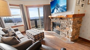 A living room with a stone fireplace, leather sofas, a patterned ottoman, and a large TV on the wall.