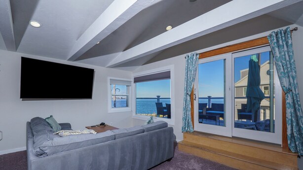 Ein Wohnzimmer mit grauem Sofa, einem Flachbildfernseher und Meerblick durch große Fenster.