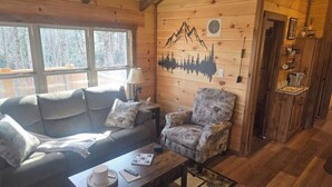 L’intérieur chaleureux d’un chalet, avec un revêtement mural en bois, un canapé, un fauteuil, une petite table sur laquelle se trouvent une tasse et un jeu, et une fresque murale représentant des montagnes et des arbres.