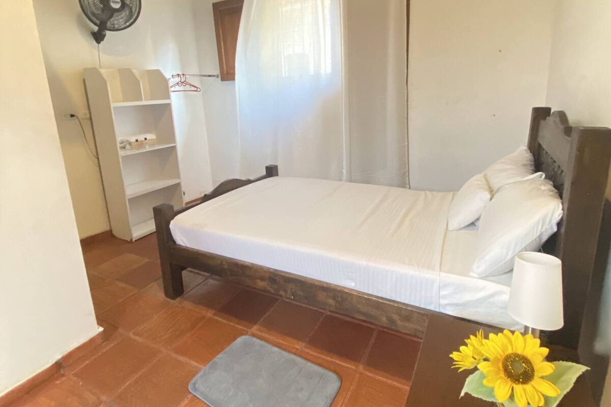 A small, clean room with a bed, a bedside table with a lamp, a fan, a shelf with toiletries, and a window with curtains.