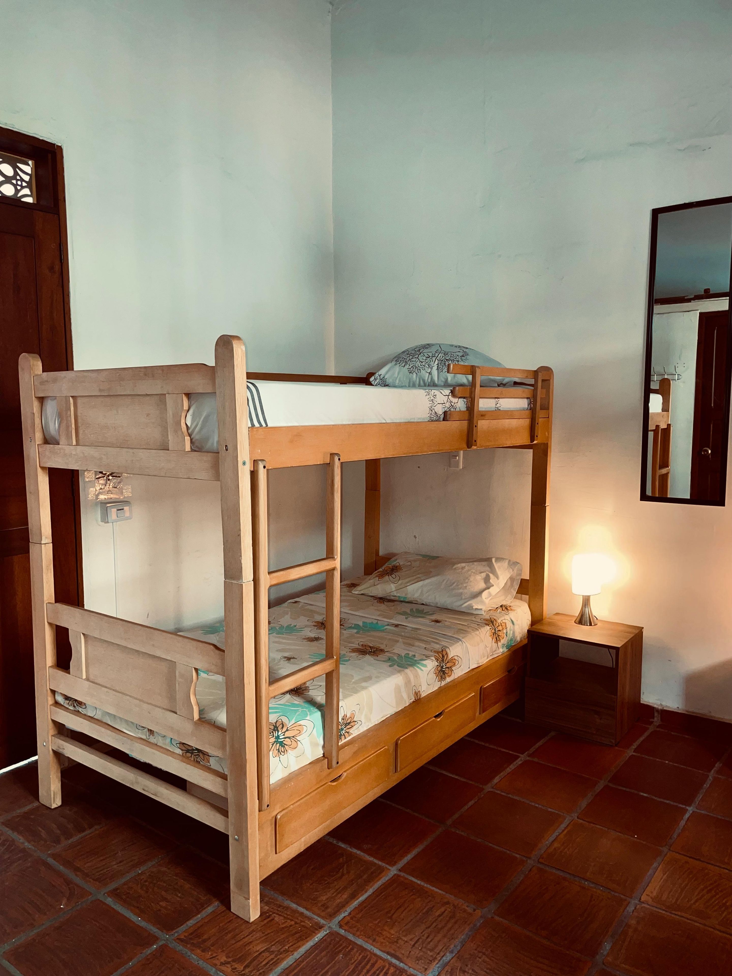 Una cama litera con estructura de madera, una mesita de noche con lámpara y un espejo en la pared.