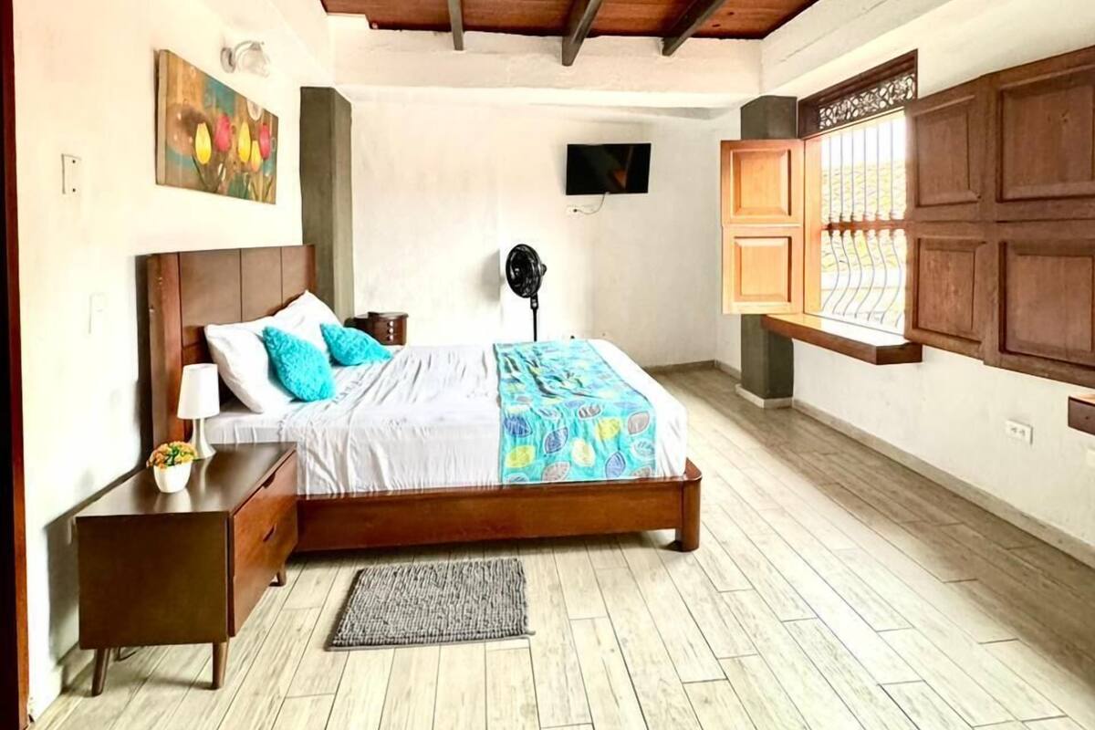A bedroom with a wooden bed, a nightstand, a bedside table, a fan, and a window with wooden shutters.