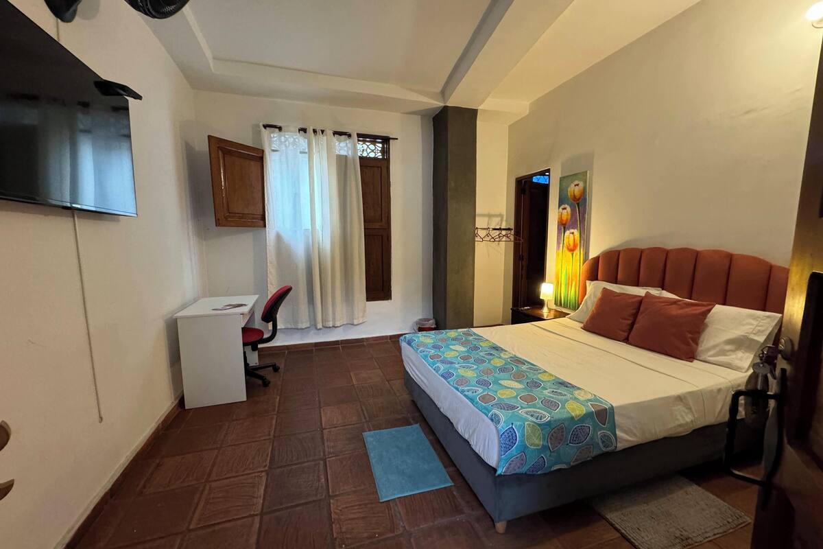 A bedroom with a bed, a desk, a chair, a TV, and a window with curtains.
