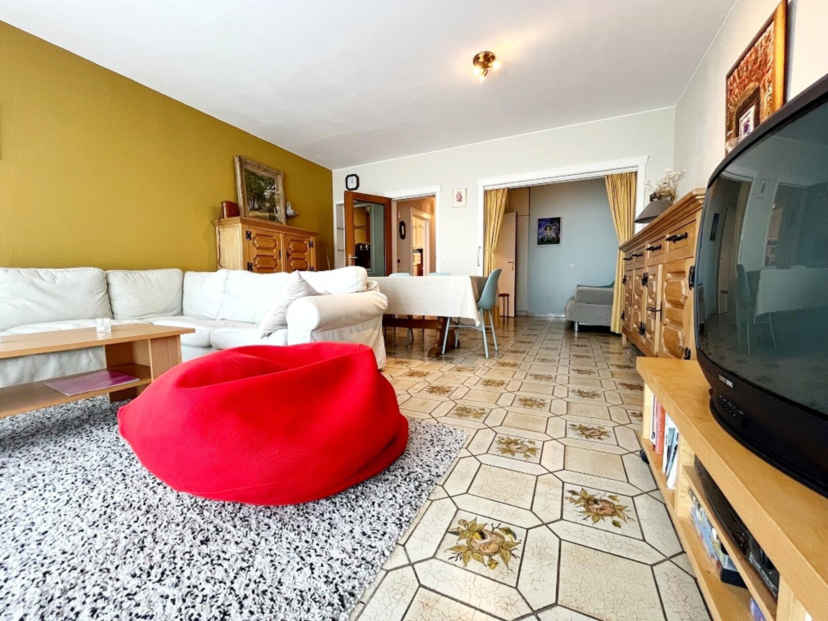A living room with a white sofa, a red bean bag, a wooden cabinet, and a television.