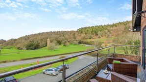 A balcony with wicker furniture, a view of a road, and a green landscape with trees. - 2 Bed in West Quantoxhead (oc-p30893) (West Quantoxhead)
