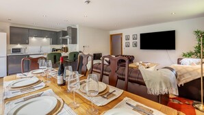 A modern living room with a dining area, a kitchen in the background, and a flat-screen TV mounted on the wall. - 2 Bed in West Quantoxhead (oc-p30893) (West Quantoxhead)