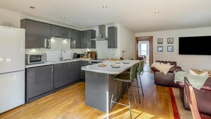 A modern kitchen with a central island, grey cabinets, and a dining area with a sofa and TV. - 2 Bed in West Quantoxhead (oc-p30893) (West Quantoxhead)