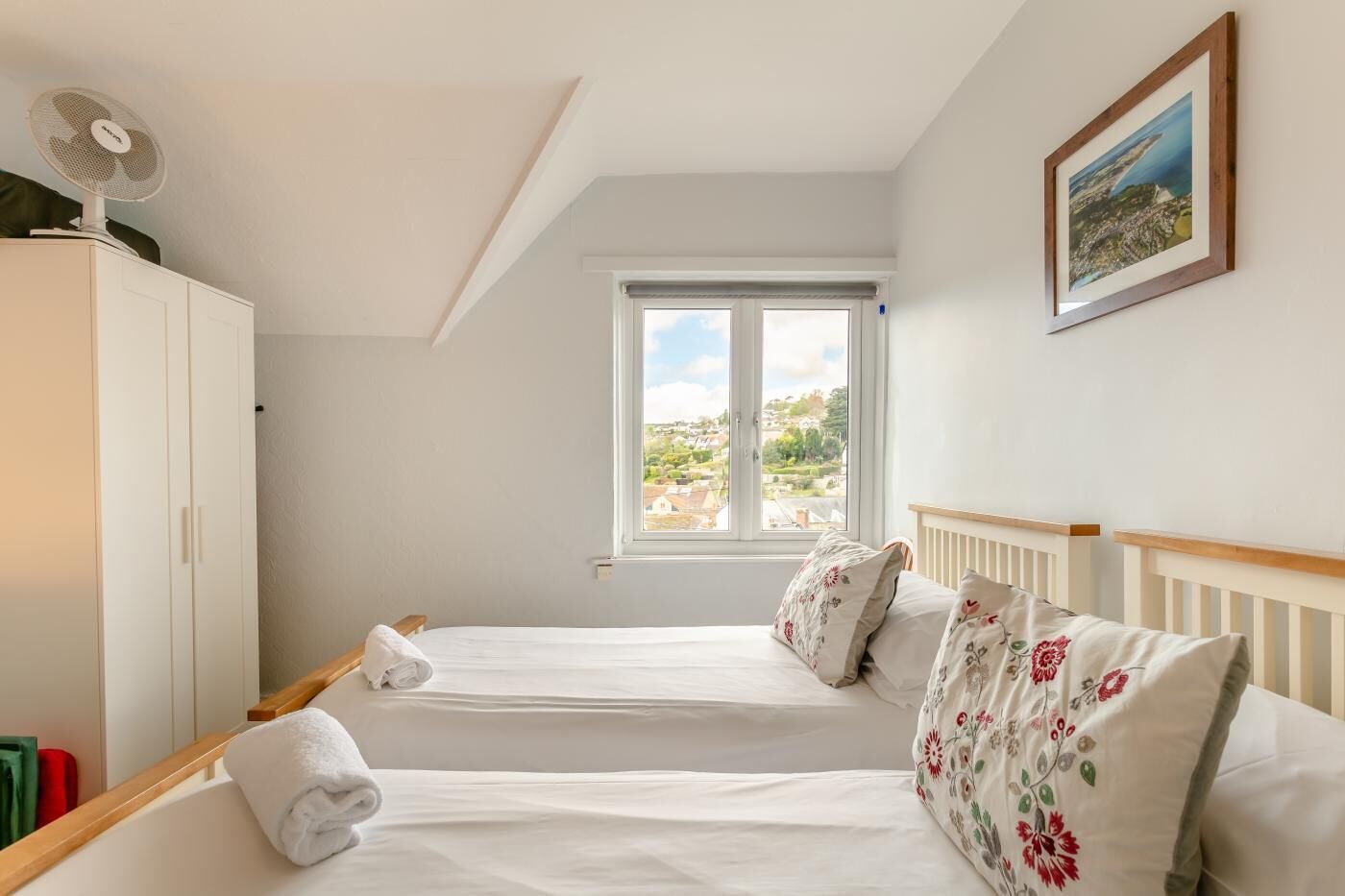A bedroom with two beds, a window with a view, a fan, and a framed picture on the wall.