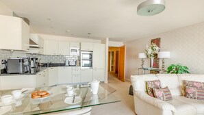 A modern kitchen with white cabinets and stainless steel appliances, a glass dining table with white chairs, and a white leather sofa with floral cushions in the living area. - 3 Bed in Torquay (oc-a31052) (Torquay)