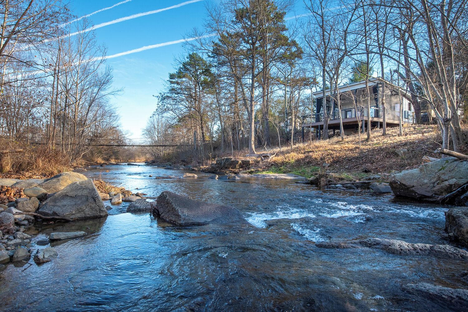 A river with rocks and trees, a bridge in the background, and a house on stilts to the right.