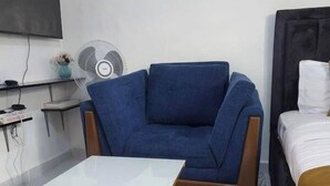 A room with a blue armchair, a white table, a bed with a dark headboard, a wall-mounted TV, and a small fan.