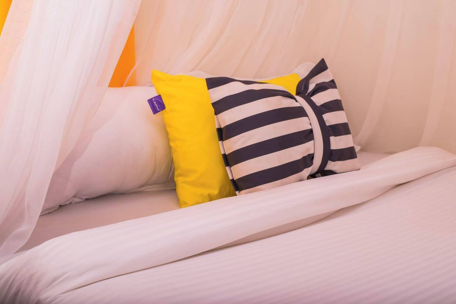 A bed with white bedding and two pillows, one striped and one solid yellow.