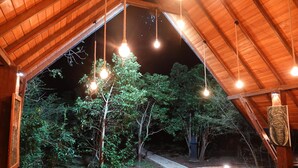 Vista interior de una estructura de madera con focos colgantes, un sendero de concreto y abundante vegetación.