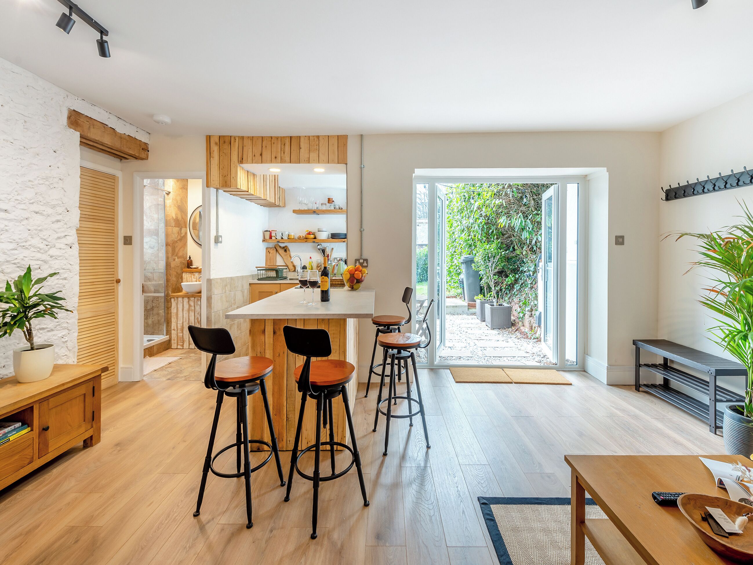 Eine moderne Küche mit Holzschränken, einer zentralen Kochinsel und Barhocker. Ein Essbereich mit Holztisch und Stühlen, sowie eine kleine Sitzecke mit einer Bank und einem Blumentopf.