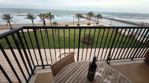 Un balcon avec vue sur l’océan, une table en bois avec une bouteille et des verres, et un espace verdoyant agrémenté de palmiers.