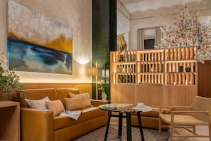 A modern hotel lobby with a large abstract painting, a wooden bookshelf, a sofa, a coffee table, and a wicker chair.
