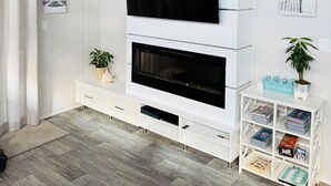 Un soggiorno moderno con una TV a schermo piatto montata su una parete bianca, un caminetto sottostante e un mobile bianco con libri e oggetti decorativi.