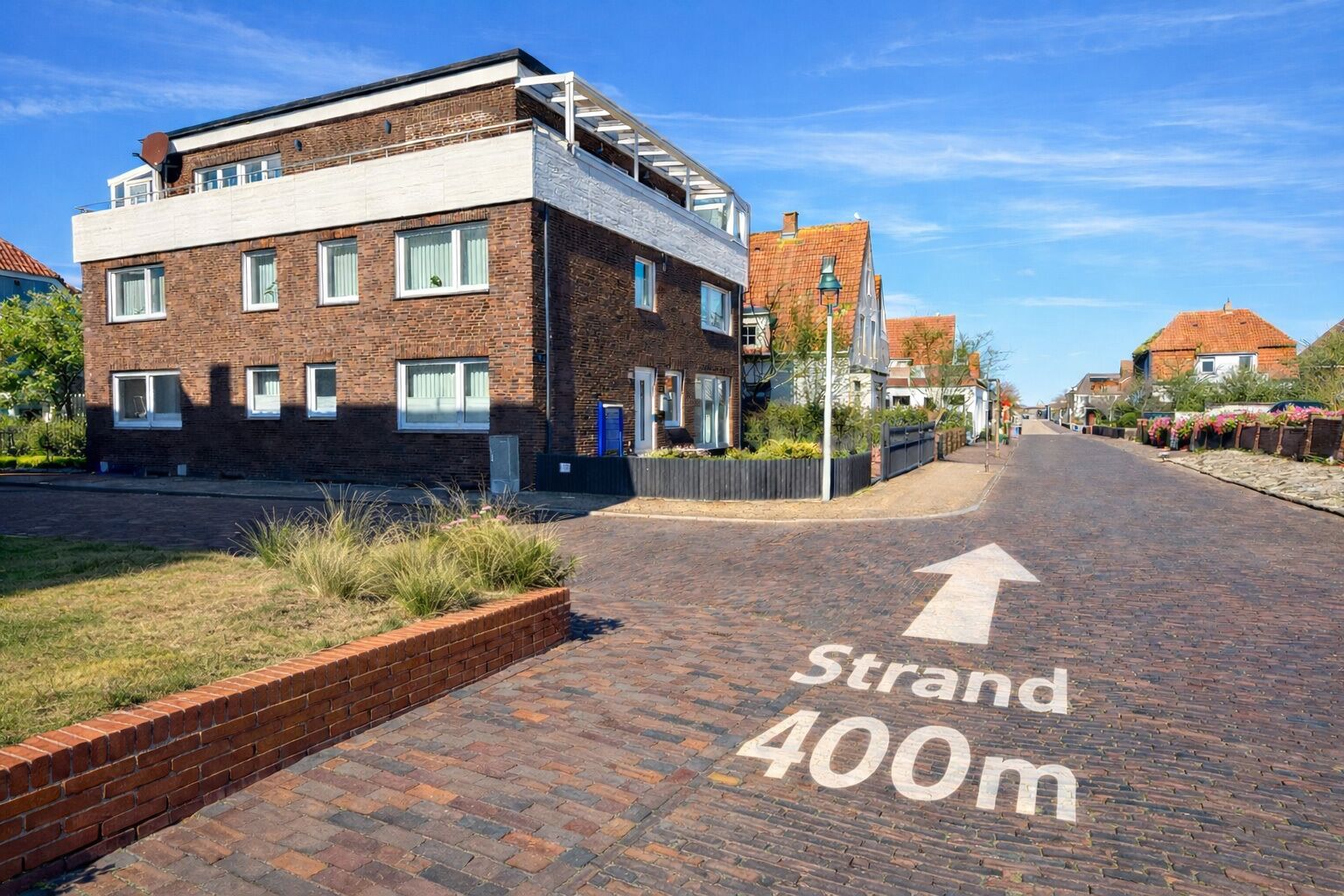 Una calle con un camino de ladrillo señalado como 'Strand 400m' y un edificio moderno con techo plano y ventanas amplias.