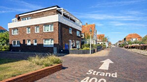 Eine Straße mit Pflasterweg, auf dem 'Strand 400m' steht, und ein modernes Gebäude mit flachem Dach und großen Fenstern.