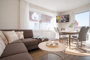 A modern living room with a sofa, a round coffee table, a dining table with chairs, and a TV mounted on the wall.