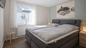 Ein Schlafzimmer mit Bett, einem Fenster mit Vorhängen, einem Heizkörper, einem kleinen Hocker und einem an der Wand befestigten Fischgemälde.