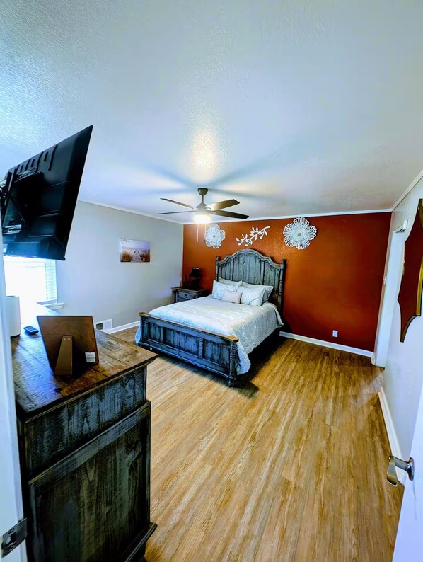 Une chambre avec un plancher en bois, un lit avec une tête de lit, un ventilateur de plafond et un téléviseur fixé au mur.